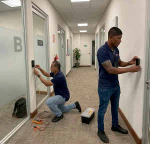 Expertos instalando sistemas de acceso de control con teclados numéricos y lectores de proximidad en pasillos de oficinas corporativas.
